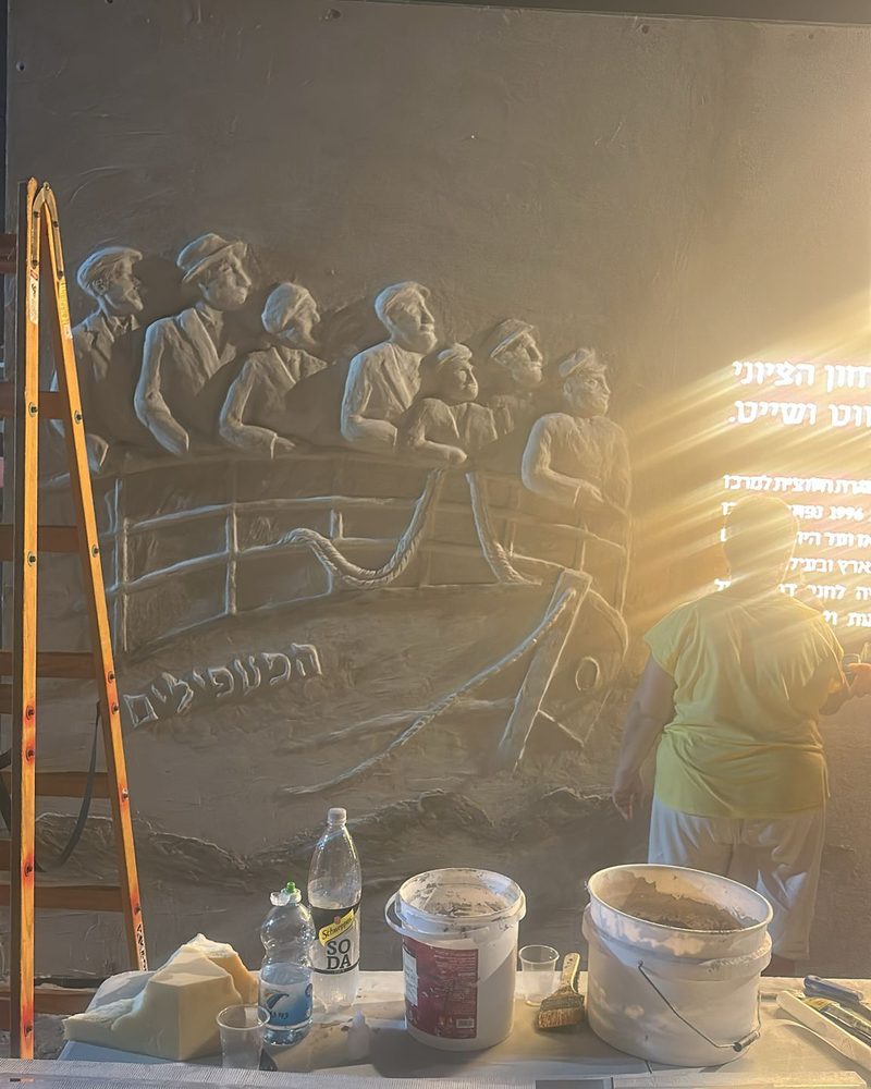 A studio member working on the large plaster relief on site, with a ladder, hand tools and buckets of plaster in low evening light