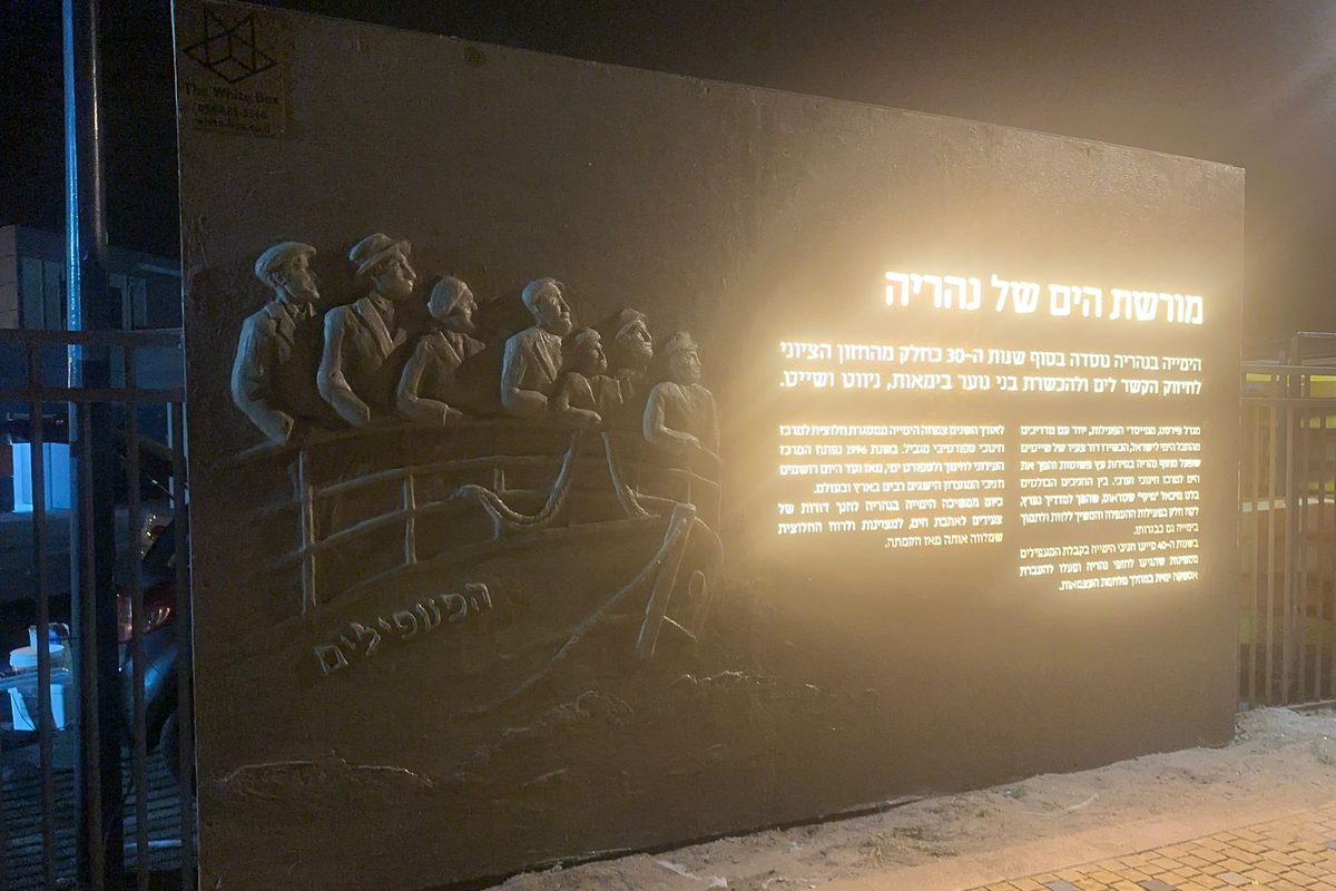 A hand-sculpted wall relief of the Ma&rsquo;apilim immigrant boat and its passengers, lit at night beside an illuminated Hebrew text panel